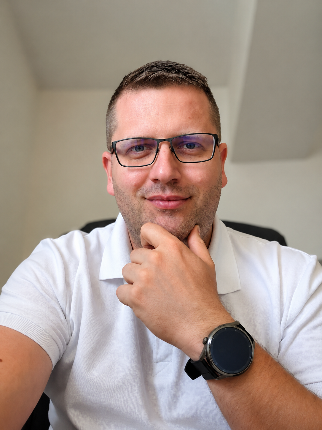 Man with glasses in a white polo shirt, resting his chin on his hand indoors.